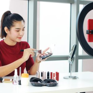 Beautiful Asian woman blogger hand holding Eye shadow box and recording on red smartphone for make up tutorial video in Cosmetics website. Marketing in social media concept.